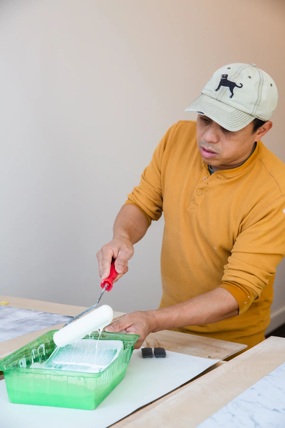 Joe, co-owner of Poppy Bee, is painting a surface with a roller and paint tray, wearing a cap and a yellow shirt.