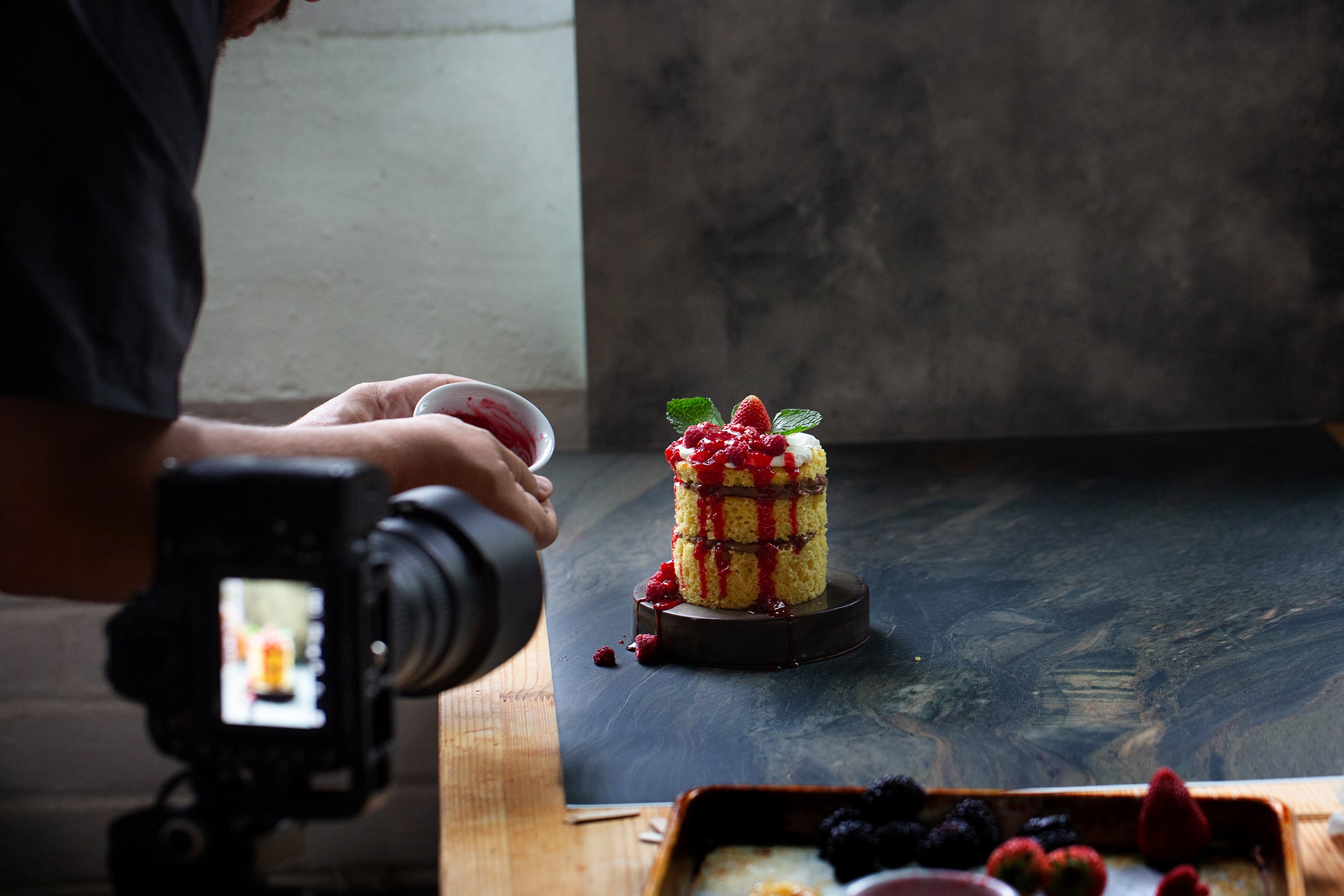 A person is taking a photo of a layered cake with berries using a camera on a wooden table.